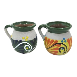 May include: Two handcrafted ceramic mugs with handles. The mugs have a gray speckled base, green rims, and colorful floral designs in yellow, orange, and blue. One mug features a blue and green design, while the other has an orange and yellow design.