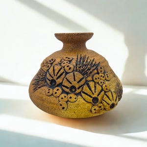 May include: A small, round, tan-colored ceramic vase with a narrow neck and a flared rim. The surface features dark, recessed, organic-looking patterns resembling sea urchins or coral clusters, with a textured, matte finish throughout the body.