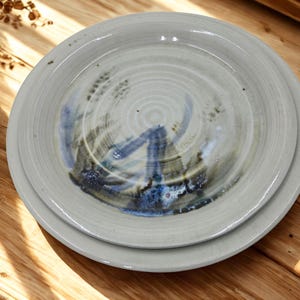 May include: A set of two round ceramic plates with a light gray, glossy finish. The plates feature a hand-painted, abstract design in shades of blue and dark green, with visible concentric circular textures and subtle dark speckles throughout.