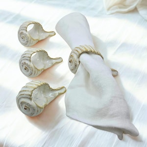 May include: A set of four ceramic napkin rings shaped like spiral seashells with a glossy off-white glaze and brown detailing along the ridges. One ring is shown holding a folded white cloth napkin, while three others are arranged separately.