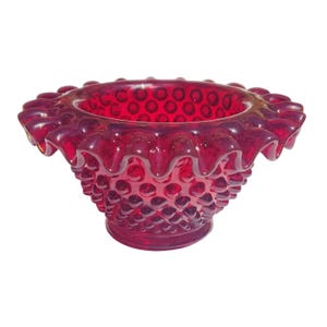 May include: A ruby red hobnail glass bowl with a ruffled rim. The bowl has a textured surface with small, rounded bumps covering the exterior. The rim is scalloped, adding a decorative touch to the vintage glassware.