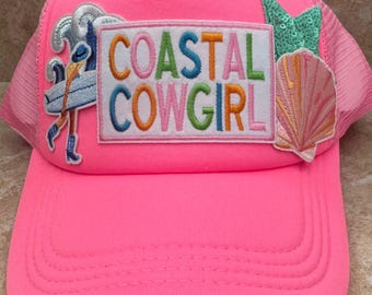 May include: Bright pink trucker hat with a mesh back. The hat features embroidered patches of a surfer, a seashell, and the words "COASTAL COWGIRL" in colorful letters. A fun, summery accessory.