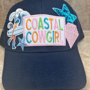 May include: Navy blue baseball cap with a mesh back and embroidered patches. The patches include a wave with a surfer, a sequined star, a pink diamond, and the words "COASTAL COWGIRL" in colorful letters.