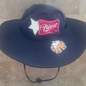 Può includere: Cappello da sole blu navy con tesa larga e sottogola. Il cappello presenta una stella bianca e una toppa rosa e bianca con la scritta "It's River Time". Sul cappello sono presenti anche due boccali di birra.