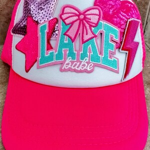 May include: A neon pink trucker hat with a white front panel. The hat features the words "LAKE babe" in turquoise and pink, along with sequined star and heart patches, and a pink lightning bolt.