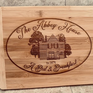 May include: A rectangular bamboo cutting board with a handle, featuring an engraved oval design. The design includes the text "The Abbey House A Bed & Breakfast" and an illustration of a house, with the establishment date "Est. 1875".