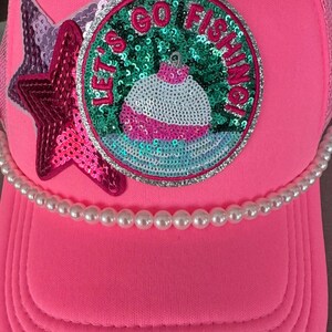 Könnte beinhalten: Hellrosa Trucker-Cap mit Perlenbesatz. Die Kappe hat einen Paillettenstern und einen runden Aufnäher mit der Aufschrift "LET'S GO FISHING" um ein Pailletten-Angelposen-Design. Die Kappe hat einen gebogenen Schirm.