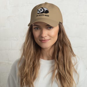 Può includere: Un cappellino da baseball color marrone chiaro con un ricamo raffigurante un panda che dorme. Sotto il panda, il testo IN SLEEPY MOOD è ricamato in lettere nere. Il cappellino presenta una visiera curva e una corona strutturata con un bottone.