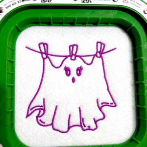 May include: A white fabric square held in a green embroidery hoop featuring a purple machine-embroidered outline of a ghost hanging from a clothesline by three clothespins. The ghost has large eyes with eyelashes and a small mouth.