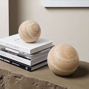Könnte beinhalten: Zwei dekorative Steinkugeln mit einem beige-braunen Marmormuster. Eine Kugel liegt auf einem Stapel Bücher mit Titeln wie "Arts the Crafts" und "House Beautiful". Die andere Kugel liegt auf einer Steinoberfläche.