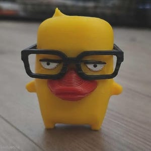 Puede incluir: Una pequeña figura amarilla con forma de pato, pico rojo y gafas rectangulares negras. La figura tiene un cuerpo liso y cuadrado con una pequeña punta en la parte superior de la cabeza y ojos blancos con pupilas negras mirando al frente.