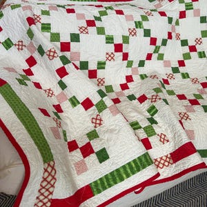 May include: A white quilt with a repeating pattern of red and green squares and crosshatch designs. The quilt has a red border and green stripes. The quilt is folded over, showing the intricate stitching and design.