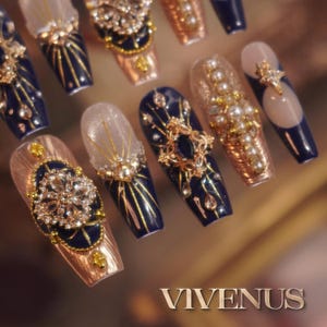 May include: A selection of long, decorated artificial nails. The nails have a dark blue base with gold detailing, rhinestones, and pearl-like beads. Some nails feature a nude base with gold and crystal adornments. The word "VIVENUS" is visible at the bottom.