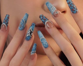 May include: Long, blue denim-style artificial nails with rhinestone accents and star embellishments. Some nails feature a brown leopard print design. The word "VIVENUS" is in the upper left corner.