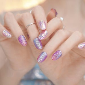 May include: Close-up of a hand with a manicure featuring iridescent purple and silver nail polish. Some nails have a swirl design, while others have glitter. A silver ring is visible on one finger.