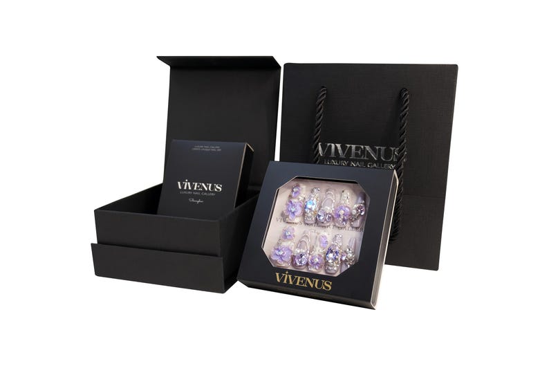 May include: Black gift set featuring artificial nails with purple flower and rhinestone embellishments. The set includes a box of nails, a small box, and a shopping bag. The text "VIVENUS Luxury Nail Gallery" is visible.