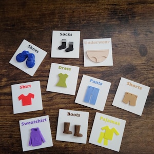 Puede incluir: Un conjunto de nueve baldosas cuadradas blancas, cada una con un icono 3D en relieve de una prenda de vestir y su etiqueta correspondiente en inglés: Shoes, Socks, Underwear, Dress, Shirt, Pants, Shorts, Sweatshirt, Boots y Pajamas.