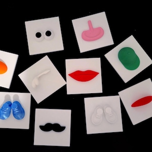 Può includere: Una collezione di piastrelle quadrate bianche, ciascuna con un elemento modellato in 3D. Gli elementi includono occhi, labbra, baffi, naso, fagiolo, scarpa, scarpa da bambino e un ovale arancione. Le piastrelle sono disposte su sfondo nero.