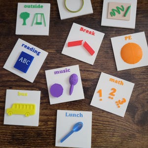 May include: A set of white square tiles with 3D-printed images and text in various colours. The tiles display words such as "Circle Time", "Outside", "Reading", "Break", "Music", "Lunch", "Math", "Art", and "PE".