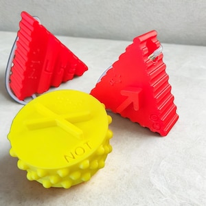 Puede incluir: Un conjunto de objetos de plástico rojos y amarillos. Dos cuñas triangulares rojas con lados estriados y un objeto redondo amarillo con la palabra "NOT" y un diseño de cruz. Los objetos parecen ser dispositivos de seguridad o de advertencia.