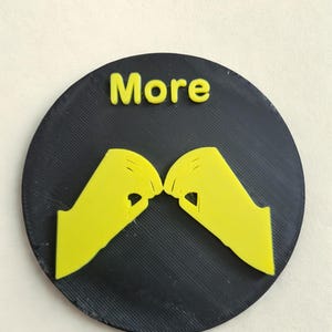 May include: A black circular object with the word "More" in yellow at the top. Two yellow hand gestures are positioned below the word, with the index fingers and thumbs touching. The object has a textured surface.