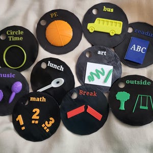 Op de afbeelding: Een verzameling zwarte, ronde tokens met verschillende thema's. Elke token toont een andere activiteit, zoals "Circle Time", "PE", "Bus", "Reading", "Art", "Music", "Lunch", "Math", "Break" en "Outside".