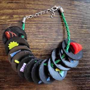 Könnte beinhalten: Ein Armband mit einer grünen geflochtenen Kordel, auf die mehrere schwarze kreisförmige Scheiben aufgefädelt sind. Jede Scheibe ist mit kleinen bunten Kunststoffanhängern verziert, darunter eine gelbe Sonne, ein roter Hut und verschiedene Formen in Blau, Lila und Orange.