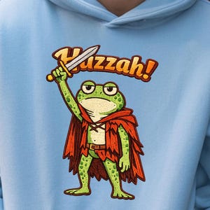 以下が含まれることがあります： 水色のパーカー。漫画のキャラクターであるカエルが剣を持ち、赤いマントを着ています。カエルの上には「Huzzah！」の文字。パーカーにはフロントポケットとドローストリングフードが付いています。