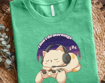 Puede incluir: Camiseta verde claro con un gato de dibujos animados durmiendo, con auriculares y un mando de videojuegos. El diseño incluye el texto "I don't do mornings" y un fondo morado y azul.