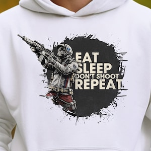May include: White hoodie featuring a graphic of a soldier holding a rifle, set against a black splatter design. The text "EAT SLEEP DON'T SHOOT REPEAT" is printed on the hoodie. The hoodie has a front pocket and a drawstring hood.