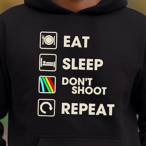May include: Black hooded sweatshirt with the words "EAT SLEEP DON'T SHOOT REPEAT" in white block letters. The text is accompanied by four icons: a plate, a bed, a target, and a recycling symbol.