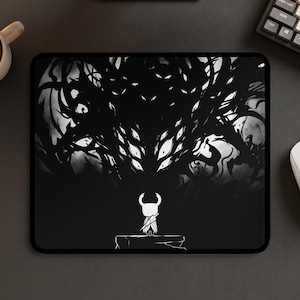 May include: A rectangular mouse pad featuring a black and white graphic illustration. A small, white, horned character stands on a ledge facing a large, dark, shadowy creature with multiple glowing white eyes in a high-contrast, monochromatic scene.