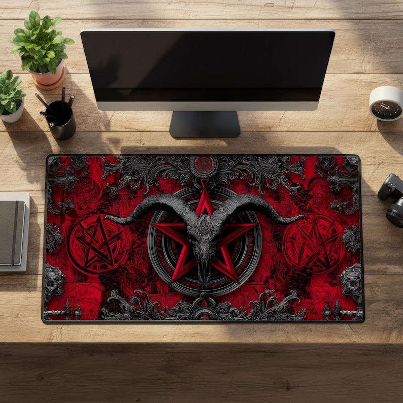 May include: A rectangular red and black desk mat featuring a central pentagram design with a goat skull, surrounded by ornate scrollwork. Two pentagrams are on either side of the central design. The mat is on a wooden desk with a computer monitor and plants.