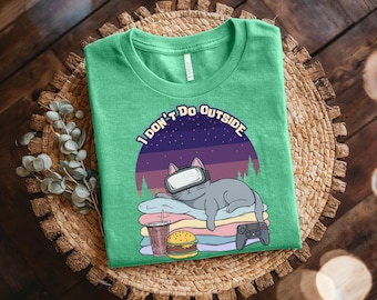 May include: A heather green t-shirt featuring a graphic of a gray cat wearing a virtual reality headset while lying on a stack of blankets. The design includes a burger, a drink, a game controller, and the text I DON'T DO OUTSIDE in white letters.