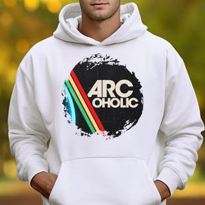 Puede incluir: Una sudadera con capucha blanca que presenta un dise&ntilde;o gr&aacute;fico central. El dise&ntilde;o consiste en una forma circular negra con textura desgastada, que contiene el texto ARC OHOLIC en blanco. Aparecen tres rayas diagonales en rojo, amarillo y verde.