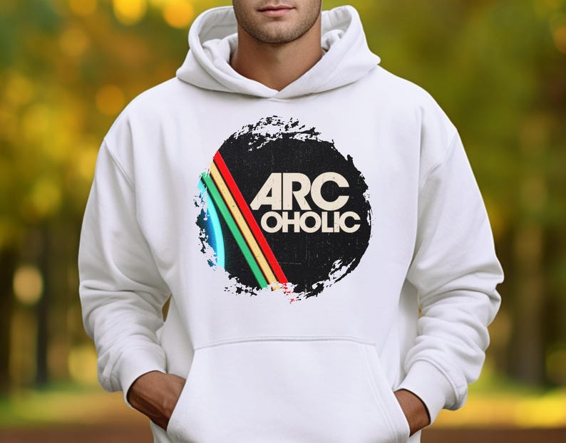 Puede incluir: Una sudadera con capucha blanca que presenta un dise&ntilde;o gr&aacute;fico central. El dise&ntilde;o consiste en una forma circular negra con textura desgastada, que contiene el texto ARC OHOLIC en blanco. Aparecen tres rayas diagonales en rojo, amarillo y verde.