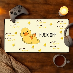 May include: A rectangular mousepad with a yellow background featuring a cartoon duck and the words "FUCK OFF" in black. The design includes yellow duck footprints and brown wavy lines. A gray game controller and headphones are visible.