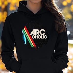 Puede incluir: Una sudadera con capucha negra que presenta un gr&aacute;fico circular en el pecho. El dise&ntilde;o incluye el texto ARC OHOLIC en letras may&uacute;sculas blancas, acompa&ntilde;ado de tres rayas diagonales en azul, verde y rojo sobre un fondo negro desgastado.