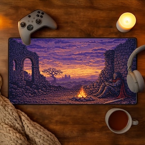 May include: A rectangular desk mat featuring a pixel art landscape of a knight in armor sitting by a campfire amidst stone ruins at sunset, with a purple and orange sky, a grey game controller, headphones, a candle, and a beige knit blanket.