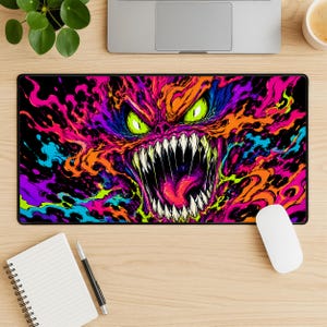 May include: A rectangular desk mat featuring a vibrant, multi-coloured graphic of a creature with glowing green eyes and sharp, pointed teeth. The design is composed of swirling patterns in shades of neon orange, purple, blue, and pink on a black background.