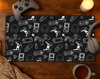 Puede incluir: Alfombrilla de ratón para juegos negra con un patrón blanco de mandos de juego, auriculares y otros accesorios de juego. Un mando gris, auriculares y una taza de café están sobre la superficie de madera. También hay una vela encendida.