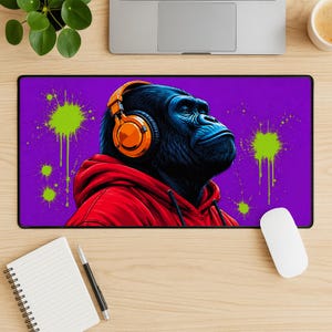 May include: A rectangular desk mat featuring a dark blue gorilla wearing bright orange headphones and a red hooded sweatshirt. The background is a vibrant purple colour with neon green paint splatter effects across the surface of the mat.