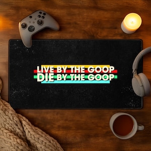May include: A rectangular black desk mat featuring the text "LIVE BY THE GOOP DIE BY THE GOOP" in white capital letters. The text is layered over horizontal stripes in yellow, red, green, and light blue, centered on the mat's surface.