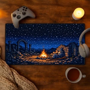 May include: A rectangular pixel art mousepad featuring a wizard sitting by a campfire under a starry night sky. The design includes ruins and a mountain range. A gray game controller, headphones, and a cup of tea are on a wooden surface.