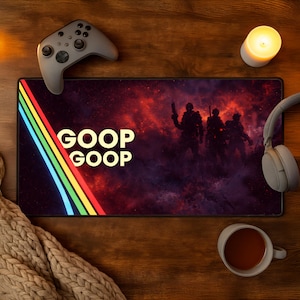 以下が含まれることがあります： 「GOOP GOOP」の文字と左側に虹色のデザインが施された長方形のゲーミングマウスパッド。パッドには、兵士のシルエットが描かれた宇宙をテーマにしたグラフィックが特徴です。グレーのコントローラー、ヘッドフォン、紅茶のカップも表示されています。