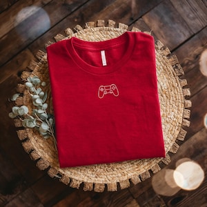 May include: A red t-shirt with a white outline of a video game controller on the front. The shirt is folded and resting on a woven, circular placemat. The background is a dark wooden surface.