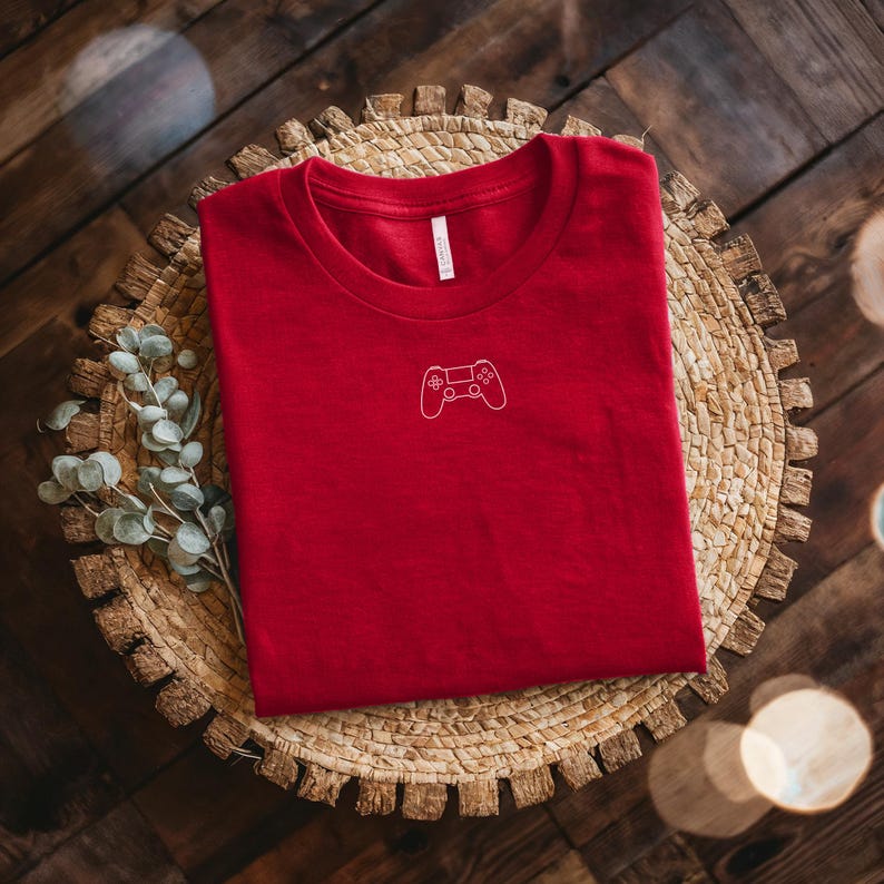 May include: A red t-shirt with a white outline of a video game controller on the front. The shirt is folded and resting on a woven, circular placemat. The background is a dark wooden surface.