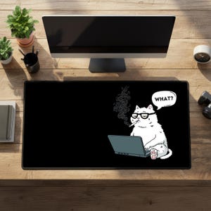 May include: Black rectangular desk mat featuring a cartoon cat with glasses, smoking a cigarette, and using a laptop. The cat has a speech bubble that says "WHAT?". The mat is on a wooden desk with a computer monitor and plants.