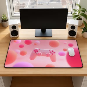 May include: A pink gaming mouse mat with a black border, featuring a pink game controller and white mouse. The mat has a pattern of pink and orange spheres on a pink background. A computer monitor and speakers are in the background.