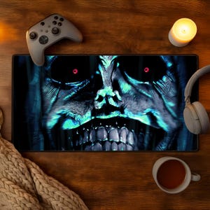 May include: A large black mousepad with a detailed, glowing blue and green monster face design. A gray game controller, headphones, a lit candle, and a mug of tea are on a wooden surface.